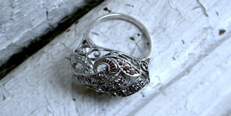 May include: A silver ring with a delicate, intricate design featuring a filigree pattern and small, clear stones. The ring is set against a white, textured background.