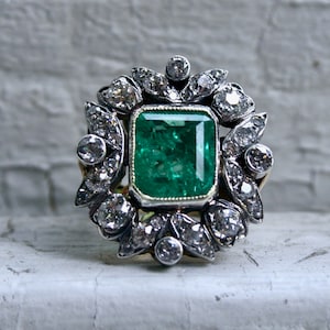 May include: A vintage emerald and diamond ring set in silver and gold. The emerald is a vibrant green and is surrounded by a halo of smaller diamonds.
