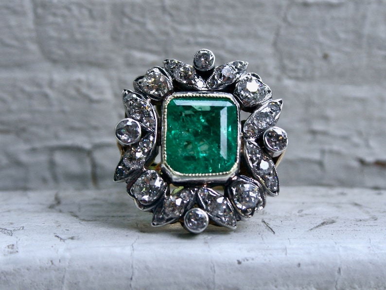 May include: A vintage emerald and diamond ring set in silver and gold. The emerald is a vibrant green and is surrounded by a halo of smaller diamonds.