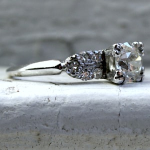 May include: A silver engagement ring featuring a large, round, clear diamond centre stone and smaller diamonds set in the band.