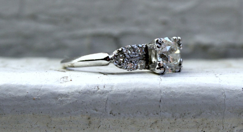 May include: A silver engagement ring featuring a large, round, clear diamond centre stone and smaller diamonds set in the band.