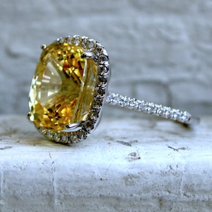 Op de afbeelding: Een close-up van een ring met een grote, ovaal geslepen gele edelsteen, omringd door een halo van kleinere, heldere stenen. De band is ook bezet met kleine, heldere stenen, wat een sprankelend effect cre&euml;ert.