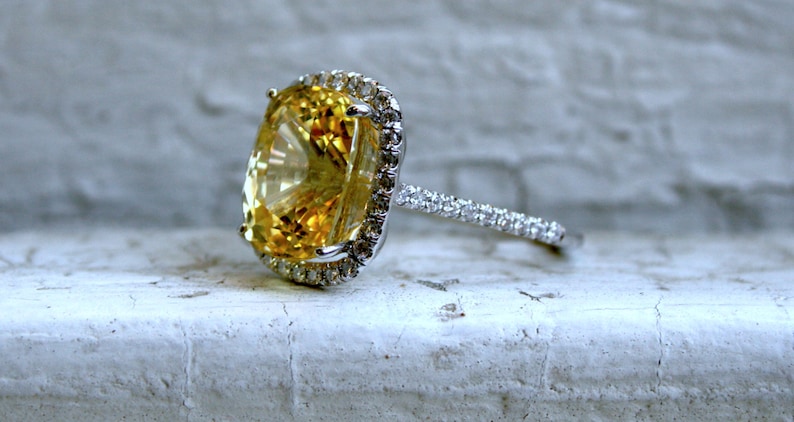 Op de afbeelding: Een close-up van een ring met een grote, ovaal geslepen gele edelsteen, omringd door een halo van kleinere, heldere stenen. De band is ook bezet met kleine, heldere stenen, wat een sprankelend effect cre&euml;ert.