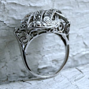 May include: A silver ring with a delicate, intricate design featuring a cluster of small diamonds. The ring has a filigree style and is set on a white background.
