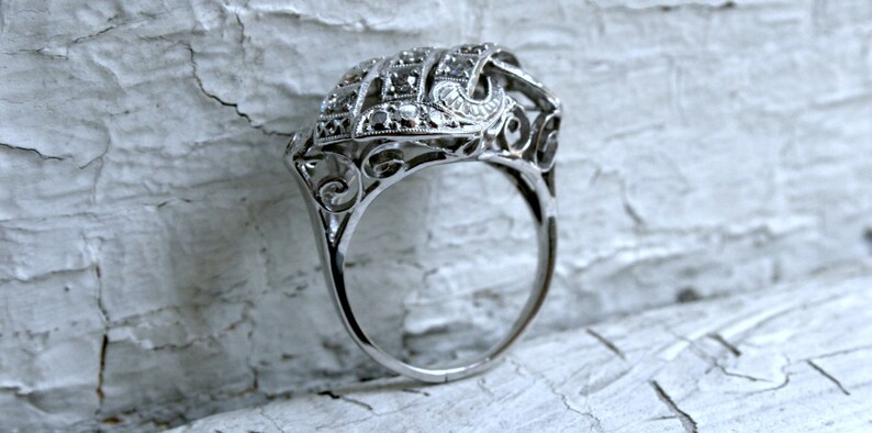May include: A silver ring with a delicate, intricate design featuring a cluster of small diamonds. The ring has a filigree style and is set on a white background.