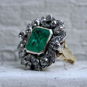 May include: A vintage emerald and diamond ring set in silver and gold. The emerald is a vibrant green and is surrounded by a halo of smaller diamonds.
