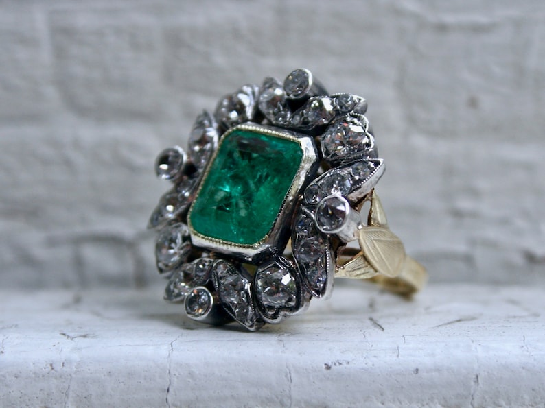 May include: A vintage emerald and diamond ring set in silver and gold. The emerald is a vibrant green and is surrounded by a halo of smaller diamonds.