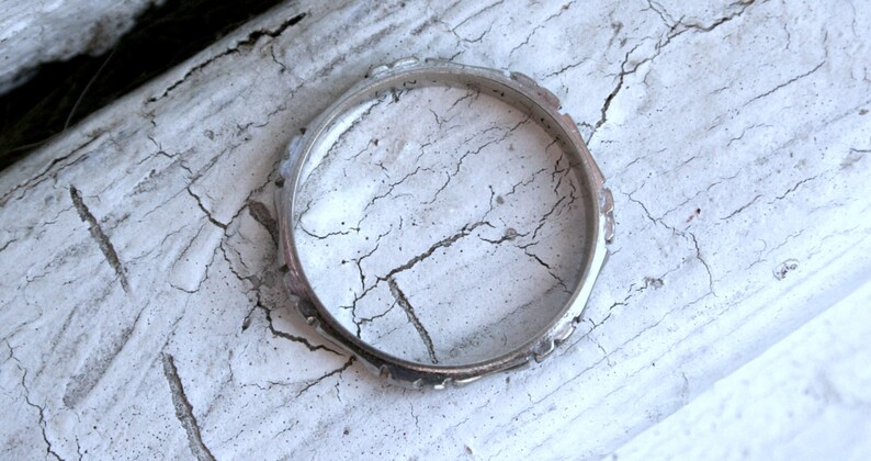 May include: A silver ring with a textured design. The ring is lying on a white, cracked surface.