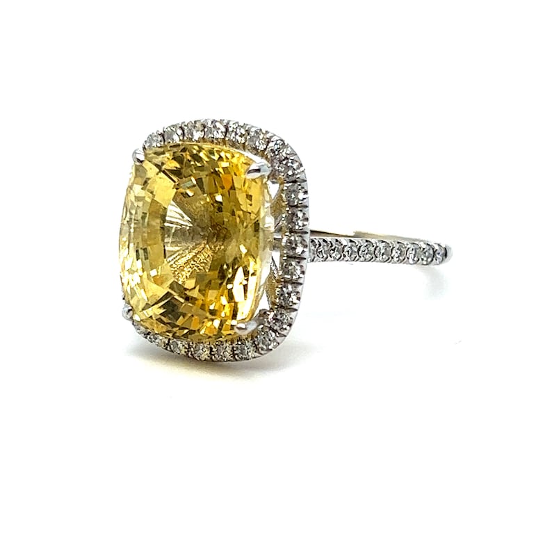 Op de afbeelding: Een opvallende ring met een grote, kussenvormige gele edelsteen, omringd door een halo van kleine, sprankelende diamanten. De ringband is ook bezet met diamanten, wat een schitterend effect cre&euml;ert. Een prachtig sieraad.