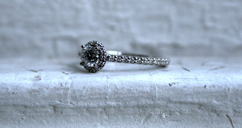 May include: A silver engagement ring with a round, clear center stone surrounded by smaller stones. The band is also set with small stones.