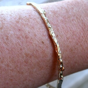 K&ouml;nnte beinhalten: Ein goldenes Armreif-Armband mit einem strukturierten, ast&auml;hnlichen Design und kleinen, klaren Steinen, die entlang der Kante gesetzt sind.