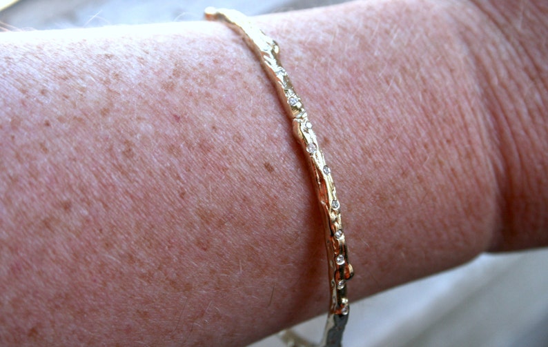 K&ouml;nnte beinhalten: Ein goldenes Armreif-Armband mit einem strukturierten, ast&auml;hnlichen Design und kleinen, klaren Steinen, die entlang der Kante gesetzt sind.