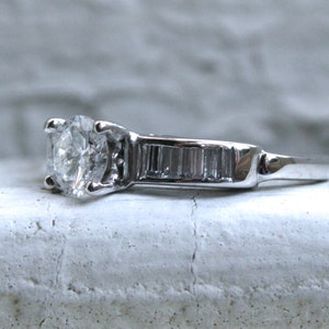 K&ouml;nnte beinhalten: Ein silberner Verlobungsring mit einem runden Diamanten in der Mitte und einer Reihe kleiner Baguette-Diamanten.