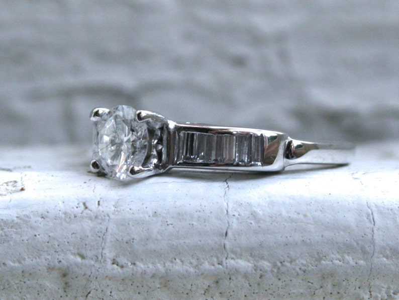K&ouml;nnte beinhalten: Ein silberner Verlobungsring mit einem runden Diamanten in der Mitte und einer Reihe kleiner Baguette-Diamanten.