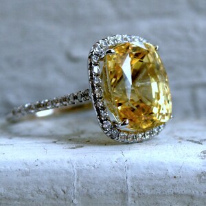 Op de afbeelding: Een zilveren ring met een grote, kussenvormige gele edelsteen. De edelsteen is omgeven door een halo van kleinere, heldere stenen. De band van de ring is ook bezet met kleine, heldere stenen. De ring wordt gepresenteerd op een wit oppervlak.