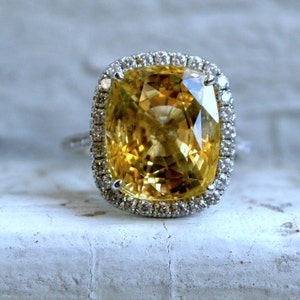 Op de afbeelding: Een ring met een grote, vierkant geslepen gele edelsteen, omgeven door een halo van kleine, heldere stenen. De band van de ring is zilverkleurig en de zetting bevindt zich op een wit oppervlak. De ring is een sieraad.