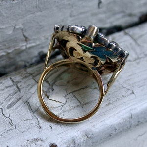 May include: A gold ring with a large, ornate setting featuring a cluster of clear and dark stones. The ring is set on a white, cracked surface.