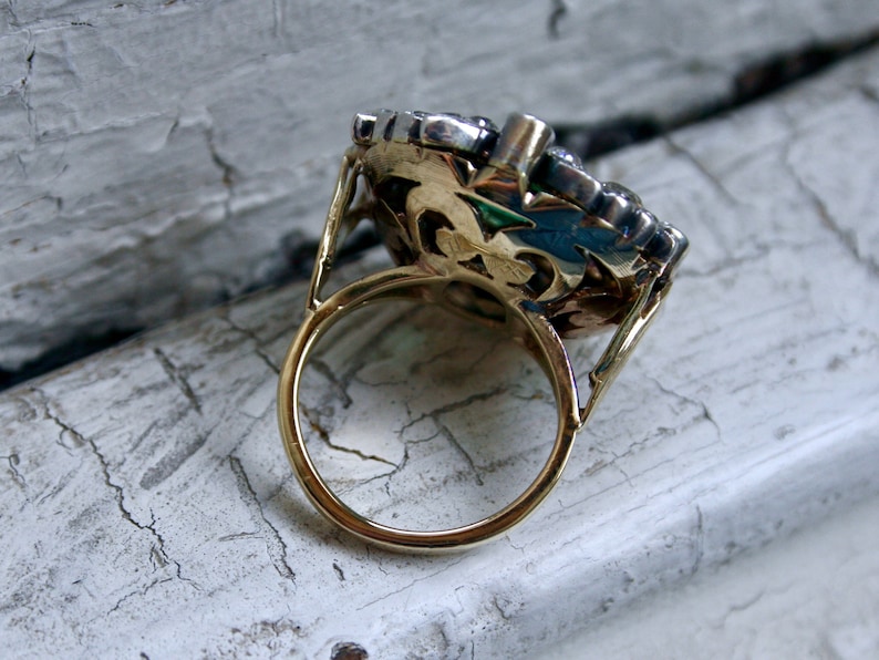 May include: A gold ring with a large, ornate setting featuring a cluster of clear and dark stones. The ring is set on a white, cracked surface.