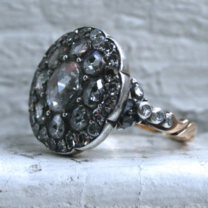 May include: A silver ring with a large oval-shaped cluster of rose-cut diamonds. The ring has a gold band.