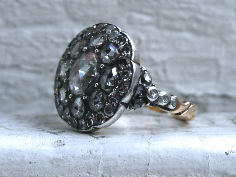 May include: A silver ring with a large oval-shaped cluster of rose-cut diamonds. The ring has a gold band.