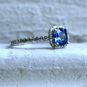 May include: A silver ring with a blue gemstone surrounded by smaller diamonds. The ring is set on a white surface.
