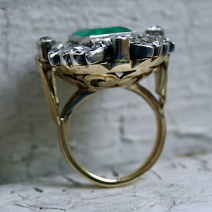May include: A gold ring featuring a large emerald green gemstone surrounded by smaller diamonds. The ring has a delicate, ornate design.