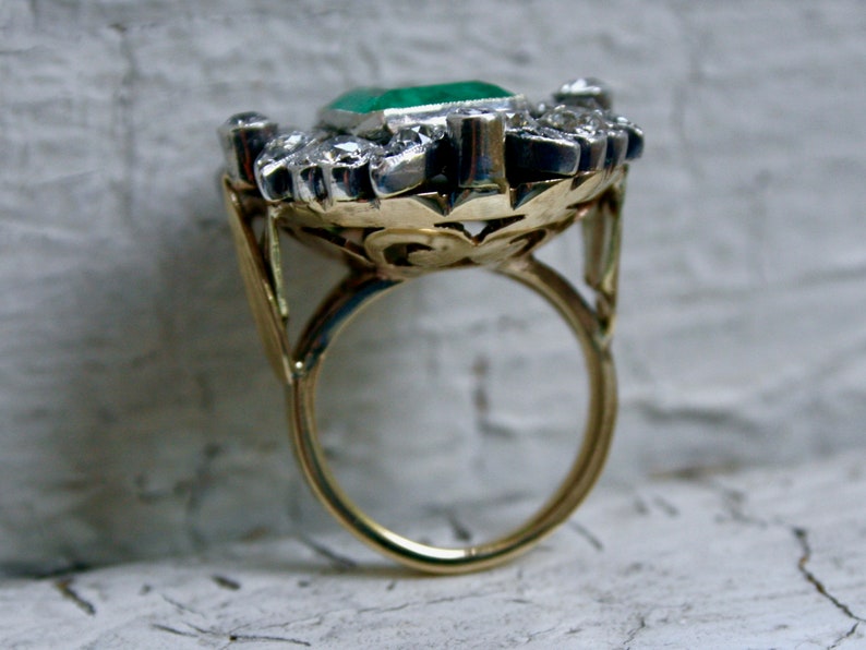 May include: A gold ring featuring a large emerald green gemstone surrounded by smaller diamonds. The ring has a delicate, ornate design.