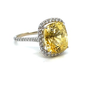 Op de afbeelding: Een opvallende ring met een grote, kussenvormige gele edelsteen, omringd door een halo van kleinere, sprankelende diamanten. De band is ook versierd met diamanten, wat een luxueus en opvallend sieraad cre&euml;ert.
