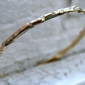 K&ouml;nnte beinhalten: Ein goldenes Armreif-Armband mit einer rauen, strukturierten Oberfl&auml;che und kleinen Diamanten, die in das Metall gefasst sind.