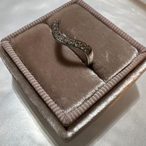 Puede incluir: Un anillo de plata con una banda curva engastada con peque&ntilde;as piedras transparentes. El anillo se muestra en una caja de anillo de terciopelo rosa claro.