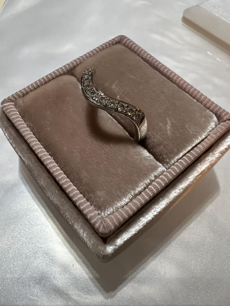 Puede incluir: Un anillo de plata con una banda curva engastada con peque&ntilde;as piedras transparentes. El anillo se muestra en una caja de anillo de terciopelo rosa claro.