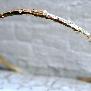 K&ouml;nnte beinhalten: Ein goldfarbenes Armreif-Armband mit einem strukturierten, ast&auml;hnlichen Design und kleinen, klaren Edelsteinen.