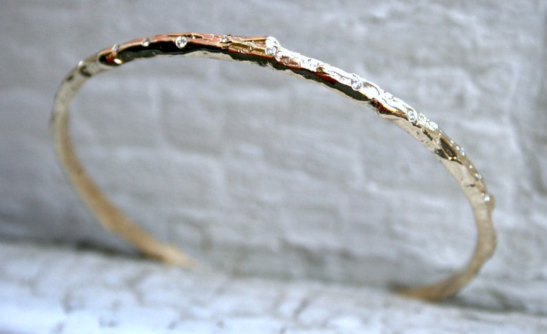 K&ouml;nnte beinhalten: Ein goldfarbenes Armreif-Armband mit einem strukturierten, ast&auml;hnlichen Design und kleinen, klaren Edelsteinen.