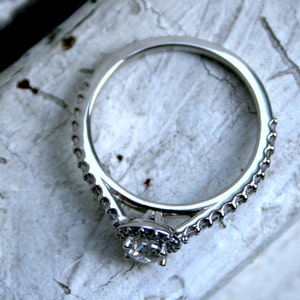 May include: A silver engagement ring with a single round diamond set in a halo setting. The ring has a delicate, pave-set band.