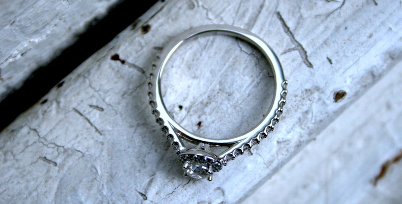 May include: A silver engagement ring with a single round diamond set in a halo setting. The ring has a delicate, pave-set band.
