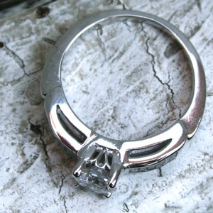 K&ouml;nnte beinhalten: Ein silberner Ring mit einem einzigen, gro&szlig;en, klaren Edelstein in der Mitte. Die Band ist mit kleineren Edelsteinen auf beiden Seiten des Mittelst&uuml;cks verziert.