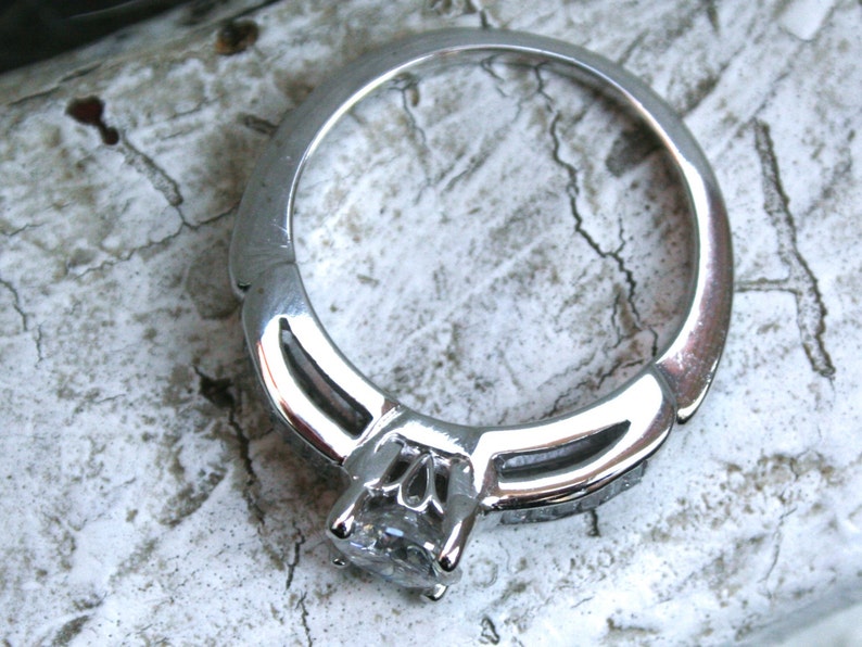 K&ouml;nnte beinhalten: Ein silberner Ring mit einem einzigen, gro&szlig;en, klaren Edelstein in der Mitte. Die Band ist mit kleineren Edelsteinen auf beiden Seiten des Mittelst&uuml;cks verziert.