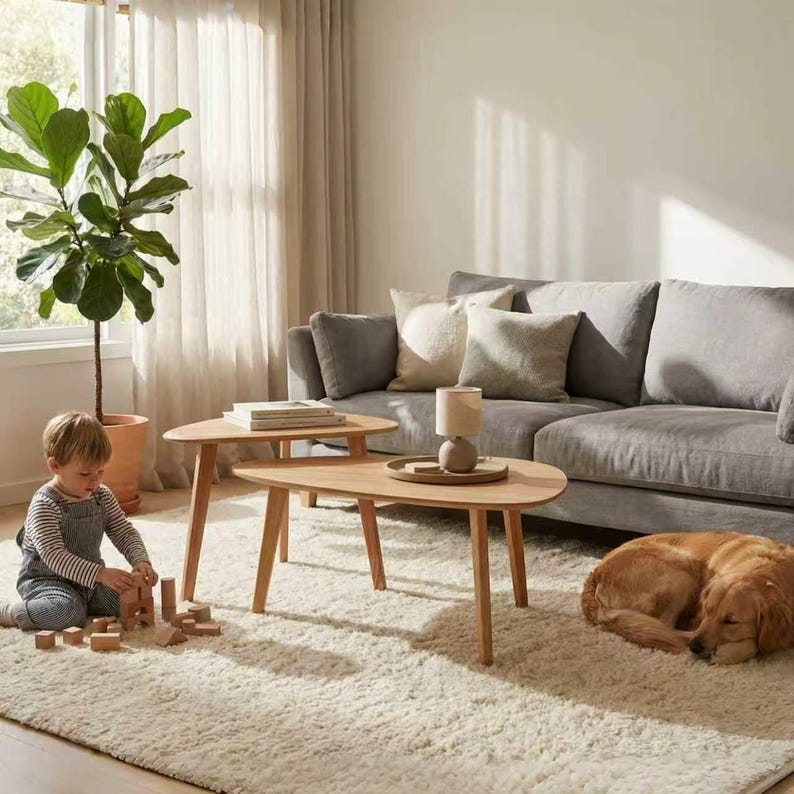 May include: A set of two light-toned wooden coffee tables with rounded triangular tops and tapered legs. The tables are arranged in a nested configuration, with a small lamp placed on the larger table, set upon a textured cream-colored rug.