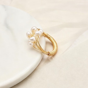 K&ouml;nnte beinhalten: Ein goldfarbener Drahtring mit einer Ansammlung kleiner, wei&szlig;er, ovaler S&uuml;&szlig;wasserperlen. Das Ringband besteht aus mehreren d&uuml;nnen, goldfarbenen Dr&auml;hten, die fest miteinander umwickelt sind und eine durchgehende, kreisf&ouml;rmige Struktur bilden.