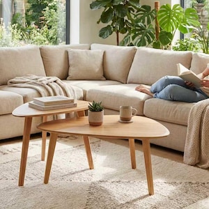 May include: Two light-toned wooden coffee tables with rounded triangular tops and three tapered legs. The tables are positioned in front of a large beige sectional sofa with matching throw pillows and a textured cream-colored area rug.