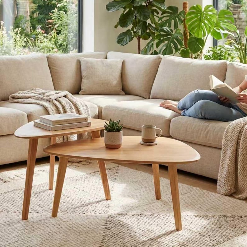May include: Two light-toned wooden coffee tables with rounded triangular tops and three tapered legs. The tables are positioned in front of a large beige sectional sofa with matching throw pillows and a textured cream-colored area rug.