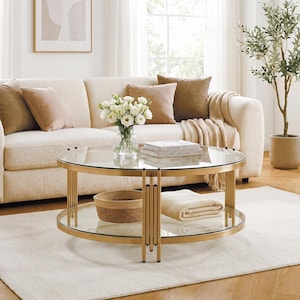 May include: A round coffee table with a gold-toned metal frame and two clear glass tiers. The table features vertical metal supports connecting the top and bottom shelves. A glass vase with white flowers and two books labeled DESIGN sit on top.