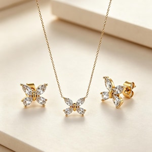 Op de afbeelding: Een goudkleurige sieradenset met een vlinderontwerp. De set bevat een ketting en een paar oorknopjes. De vlinderhangers en oorbellen zijn versierd met heldere, sprankelende edelstenen. De ketting heeft een delicate ketting.