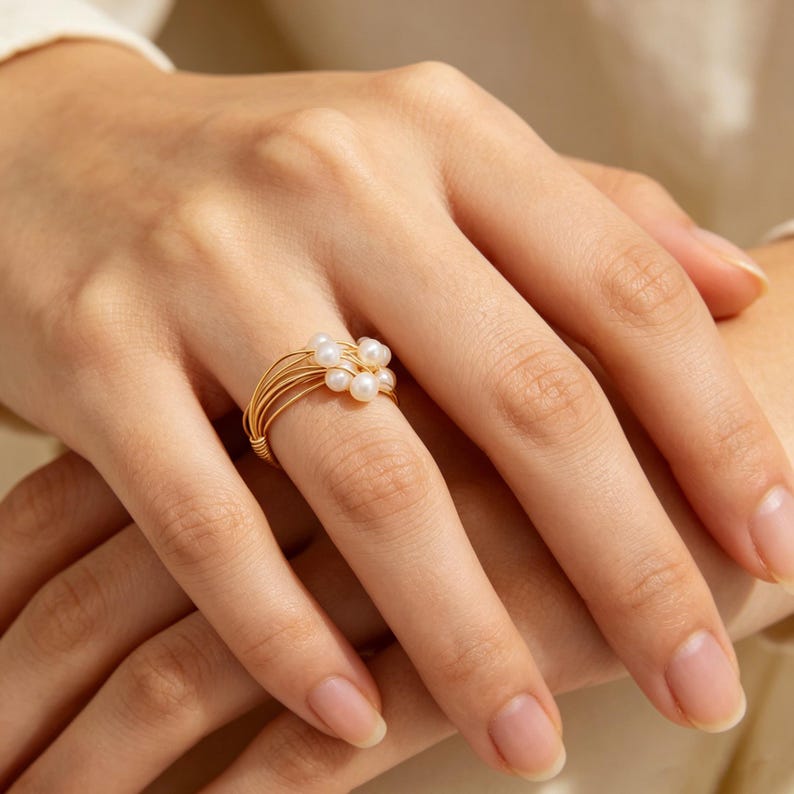 K&ouml;nnte beinhalten: Ein Ring aus goldfarbenem Draht mit einer Ansammlung kleiner, runder, wei&szlig;er Perlen. Die Ringschiene besteht aus mehreren d&uuml;nnen, parallelen goldfarbenen Dr&auml;hten, die sich um den Finger legen und zusammenlaufen, um die Perlen zu halten.