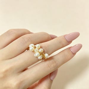 K&ouml;nnte beinhalten: Ein Ring mit mehreren kleinen, wei&szlig;en, kugelf&ouml;rmigen Perlen, die zusammen gruppiert und mit d&uuml;nnem, ineinander verschlungenem goldfarbenem Draht befestigt sind. Das Design besteht aus mehreren Perlen in einer geschichteten, gewickelten Anordnung.