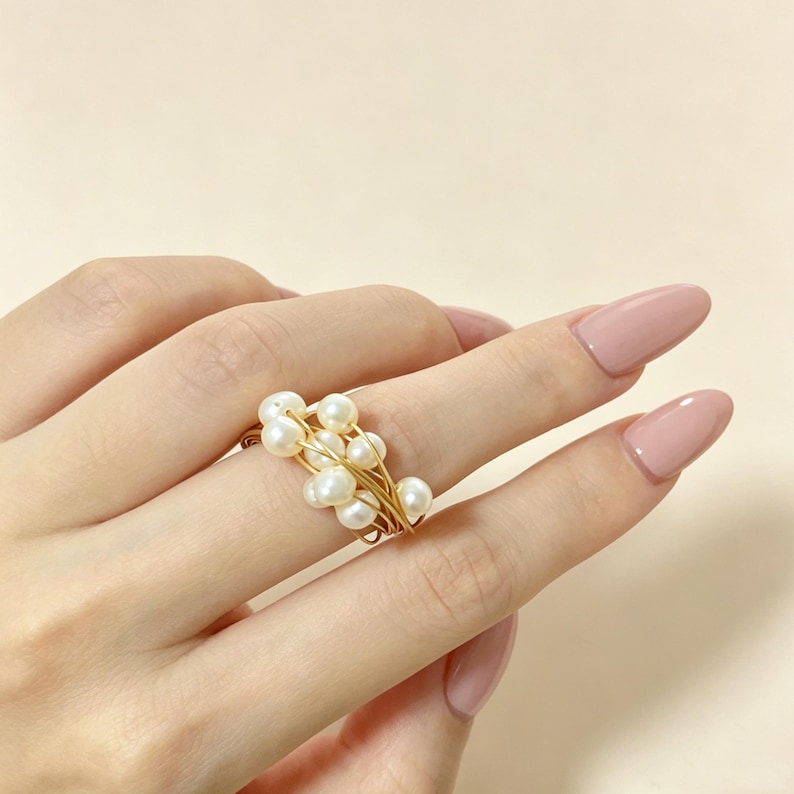 K&ouml;nnte beinhalten: Ein Ring mit mehreren kleinen, wei&szlig;en, kugelf&ouml;rmigen Perlen, die zusammen gruppiert und mit d&uuml;nnem, ineinander verschlungenem goldfarbenem Draht befestigt sind. Das Design besteht aus mehreren Perlen in einer geschichteten, gewickelten Anordnung.