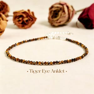 May include: An anklet featuring small, faceted, golden-brown tiger eye gemstone beads arranged in a single strand. The piece includes a silver-toned metal clasp and an adjustable extender chain. The text Tiger Eye Anklet is printed below.
