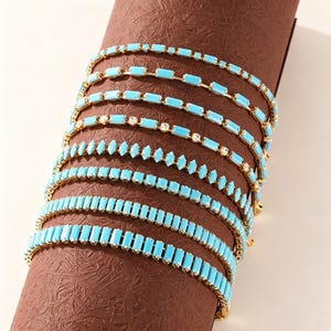 Puede incluir: Una colección de siete pulseras de tono dorado con segmentos rectangulares de color turquesa. Algunas pulseras incluyen pequeñas piedras transparentes intercaladas entre las piezas turquesas, mientras que otras presentan una fila continua.