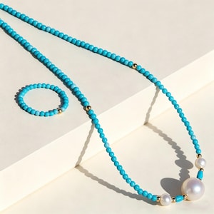 Könnte beinhalten: Ein türkisfarbenes Perlenketten- und Armband-Set mit goldenen Akzenten. Die Halskette hat einen großen weißen Perlenanhänger und kleinere Perlen. Der Schmuck wird auf einer weißen Oberfläche präsentiert.