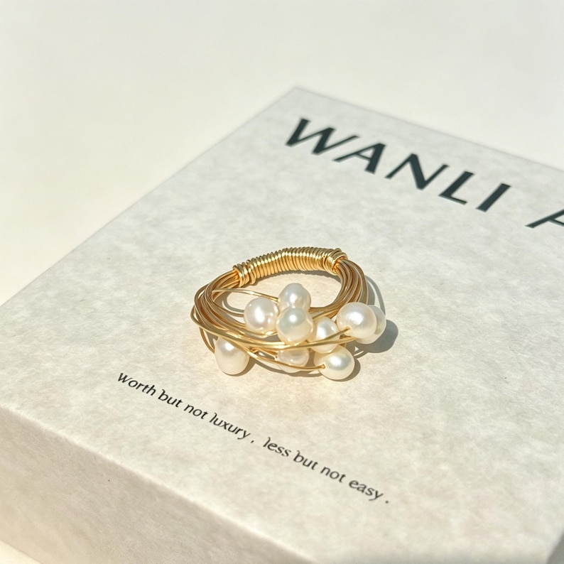 K&ouml;nnte beinhalten: Ein Ring aus mehreren Str&auml;ngen d&uuml;nnen goldfarbenen Drahtes, die um ein zentrales Band gewickelt sind, mit mehreren kleinen, runden, wei&szlig;en Perlen. Der Ring liegt auf einer wei&szlig;en Schachtel mit dem Text WANLI und Worth but not luxury, less but not easy.